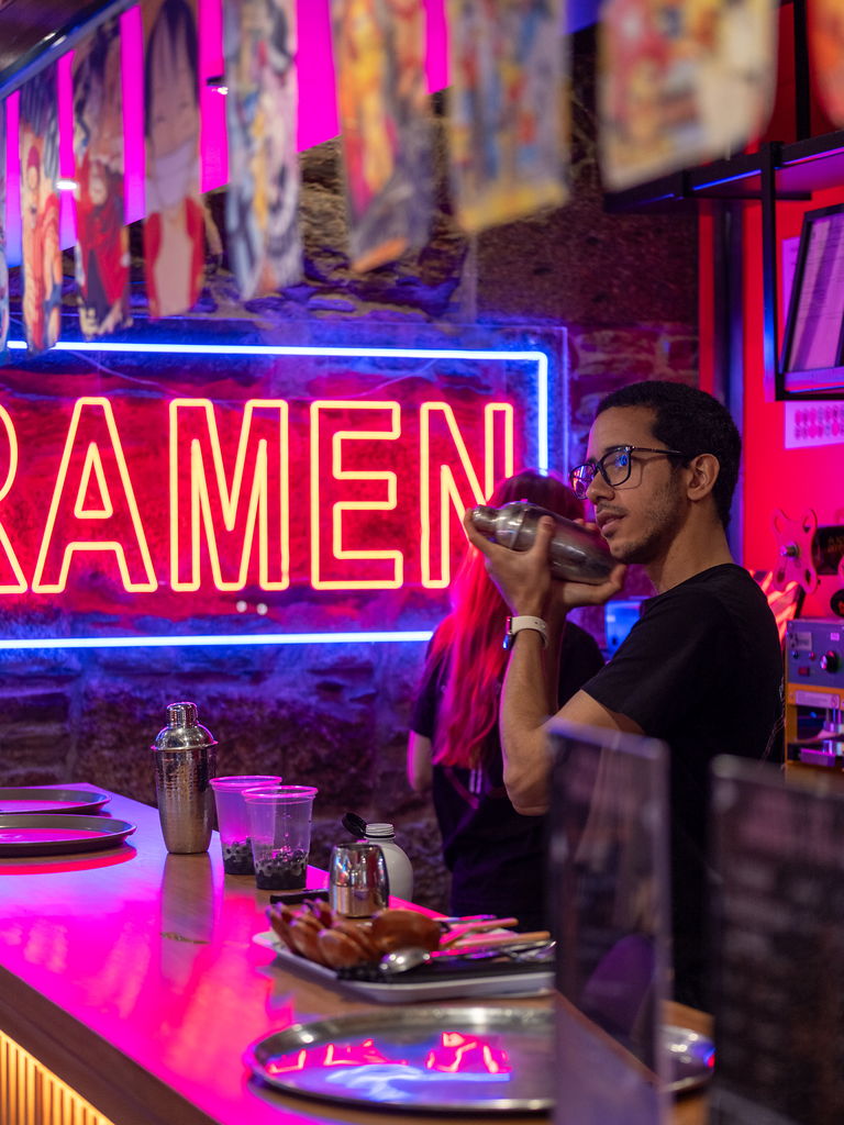 A bartender shaking a cocktail behind the bar, with the RAMEN neon behind him